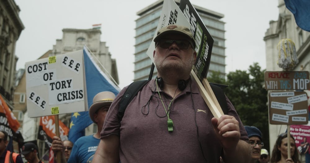 Man stands at anti-austerity protest in front of a sign reading Cost of Tory Crisis