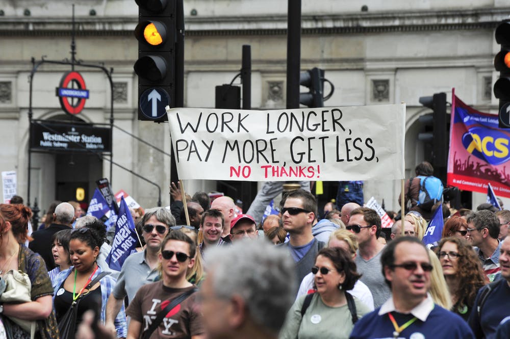 Protesters gather under a banner reading Work Longer Pay More Get Less