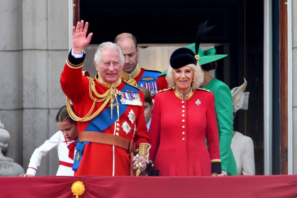 King Charles waves to a crow from the balcony at Buckingham Palace, wearing a red uniform with a blue sash and medals. Queen Camilla stands next to him wearing a red dress and black hat.