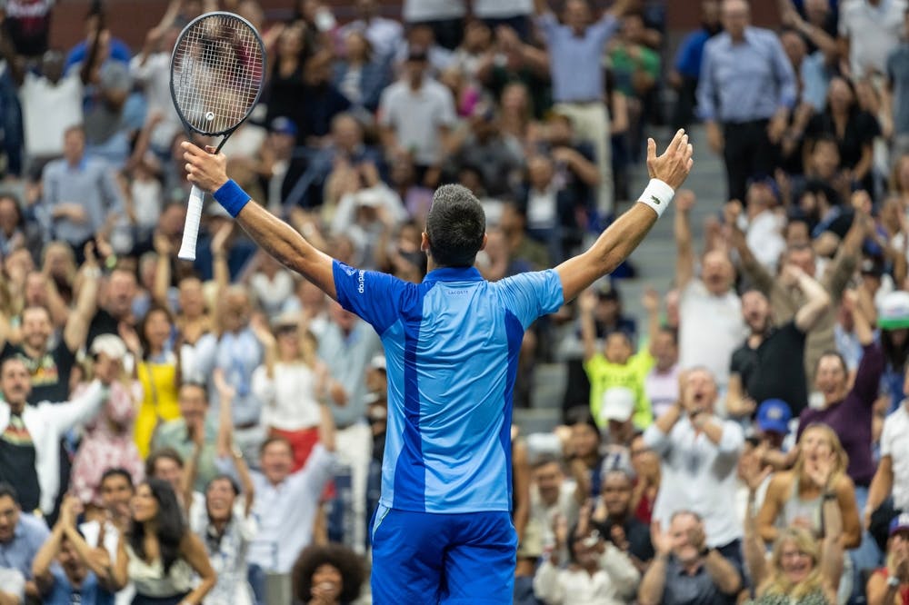 Novak Djokovic holds both arms up in celebration, facing the crowd